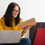 Mulher comprando online com notebook e sacolas, representando estratégia de produto de entrada no e-commerce para aumentar vendas