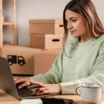 Mulher trabalhando no notebook cercada de caixas de envio, representando gestão de produtos para vender online