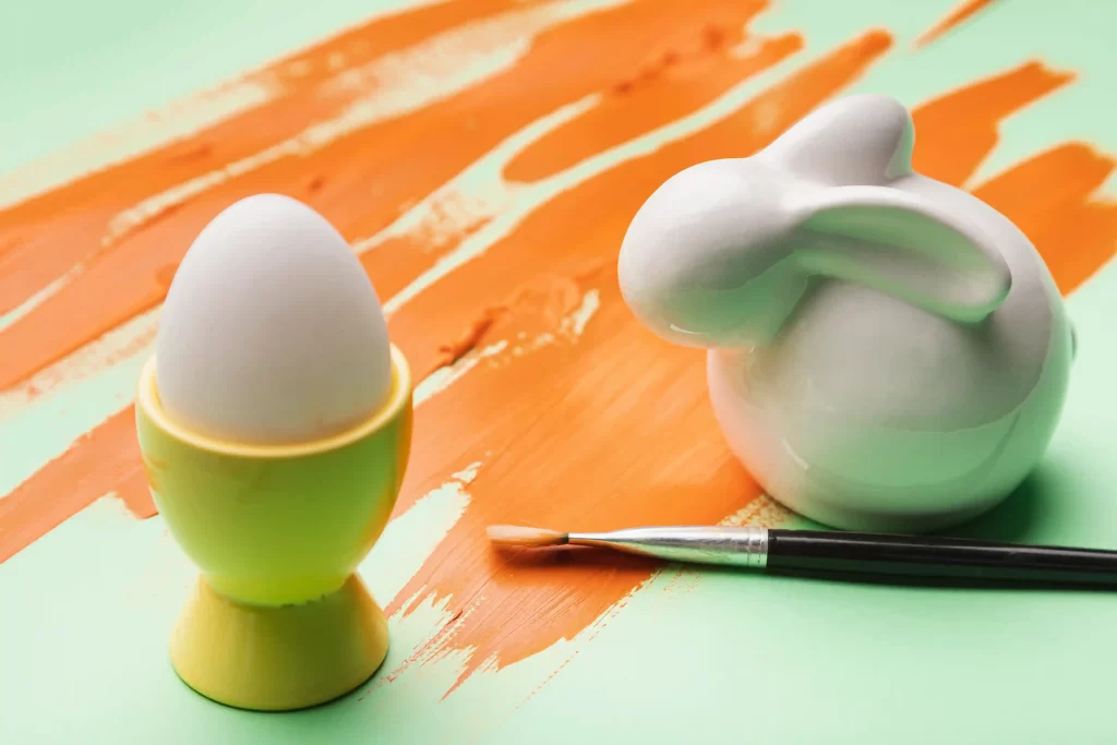 Ovo branco em suporte com pincel e objeto decorativo de coelho sobre fundo colorido, representando produtos criativos e artesanais relacionados às tendências de Páscoa 2026.
