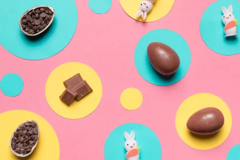 Ovos de chocolate, pedaços de chocolate e pequenos coelhos decorativos sobre fundo colorido, representando tendências e microtendências de produtos para Páscoa 2026