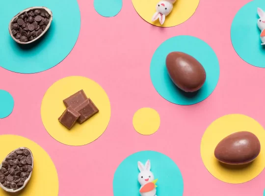 Ovos de chocolate, pedaços de chocolate e pequenos coelhos decorativos sobre fundo colorido, representando tendências e microtendências de produtos para Páscoa 2026