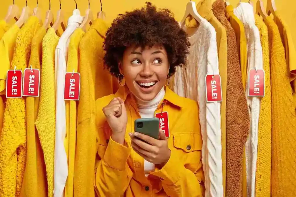 Pessoa animada em frente a arara de roupas com etiquetas vermelhas de promoção, segurando smartphone