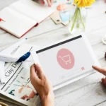 Pessoa segurando tablet com ícone de carrinho de compras na tela, em mesa com objetos de planejamento e viagem