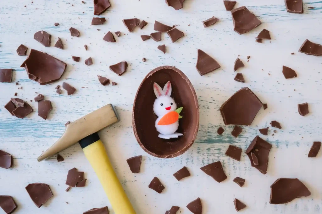 Ovo de chocolate quebrado sobre mesa branca com coelho decorativo dentro e pedaços espalhados ao redor