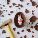 Ovo de chocolate quebrado sobre mesa branca com coelho decorativo dentro e pedaços espalhados ao redor