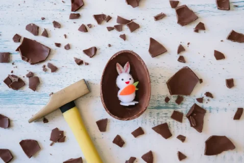 Ovo de chocolate quebrado sobre mesa branca com coelho decorativo dentro e pedaços espalhados ao redor
