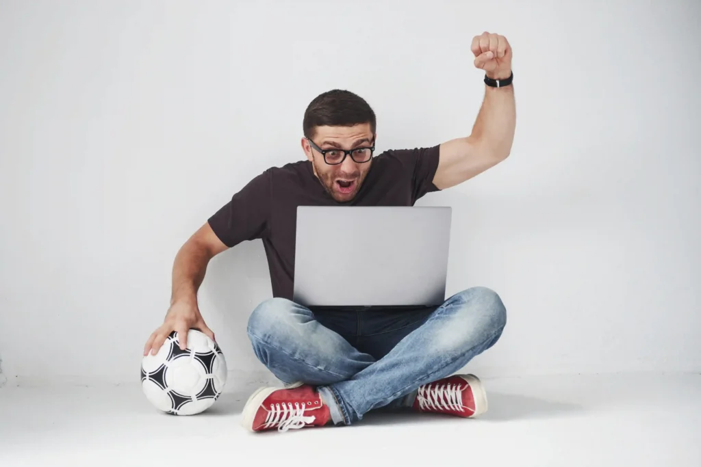 Homem empolgado com bola de futebol e notebook, representando o consumo digital durante a Copa do Mundo e o comportamento do comprador nos dias de jogo