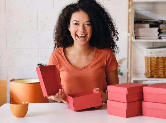 Empreendedora animada organizando caixas de produto rosa, representando a preparação do estoque dia das mães 2026