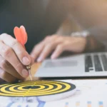 Mão segurando dardo vermelho apontado para alvo em mesa com laptop e gráficos, simbolizando foco e estratégia
