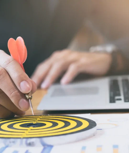 Mão segurando dardo vermelho apontado para alvo em mesa com laptop e gráficos, simbolizando foco e estratégia