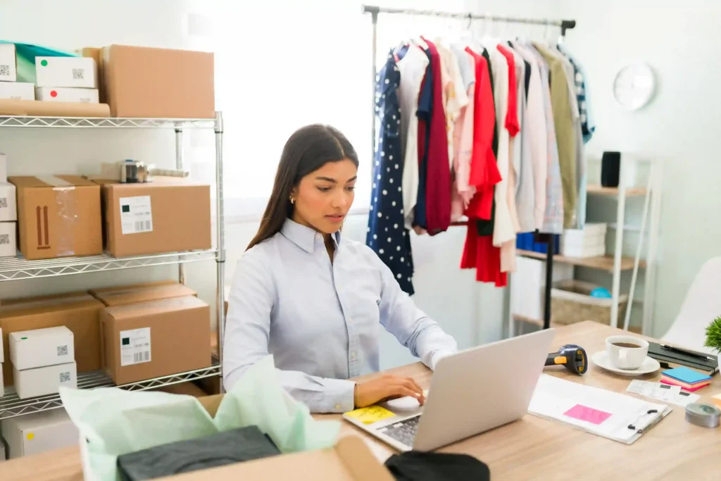 Empreendedora gerenciando loja no notebook com estoque e roupas ao fundo, representando a rotina de gestão da loja no Mercado Livre