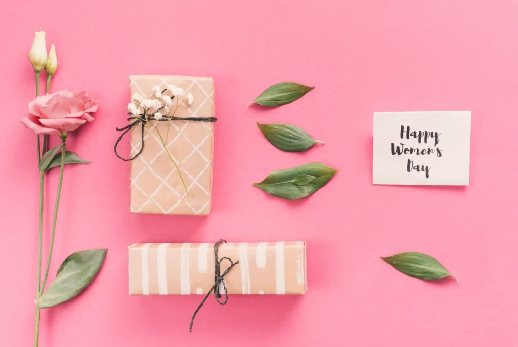 Dois presentes embrulhados em papel kraft com laços, rosa e cartão escrito Happy Women's Day sobre fundo rosa, representando a montagem de kit dia das mães para vender no Mercado Livre