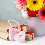 Vaso com flores coloridas ao lado de caixas de presente decoradas, simbolizando itens com apelo emocional para o Dia das Mães