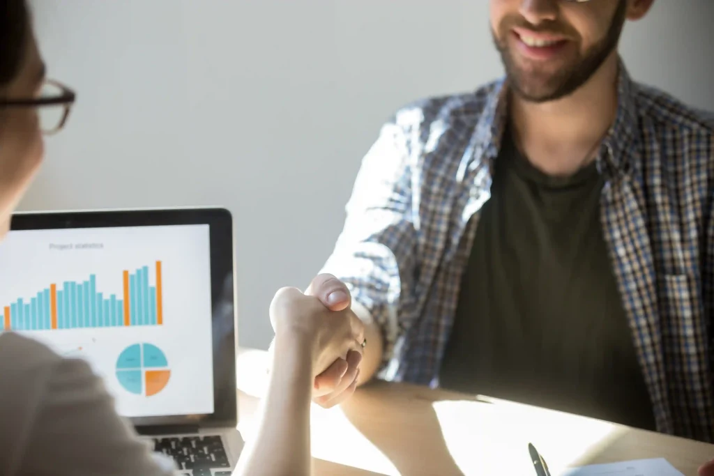 Duas pessoas em reunião apertando as mãos diante de laptop com gráficos coloridos, representando análise de tendências de mercado