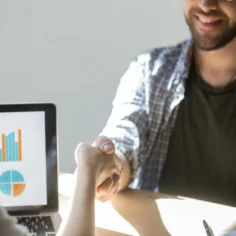Duas pessoas em reunião apertando as mãos diante de laptop com gráficos coloridos, representando análise de tendências de mercado