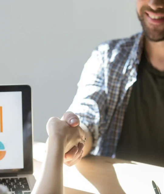 Duas pessoas em reunião apertando as mãos diante de laptop com gráficos coloridos, representando análise de tendências de mercado