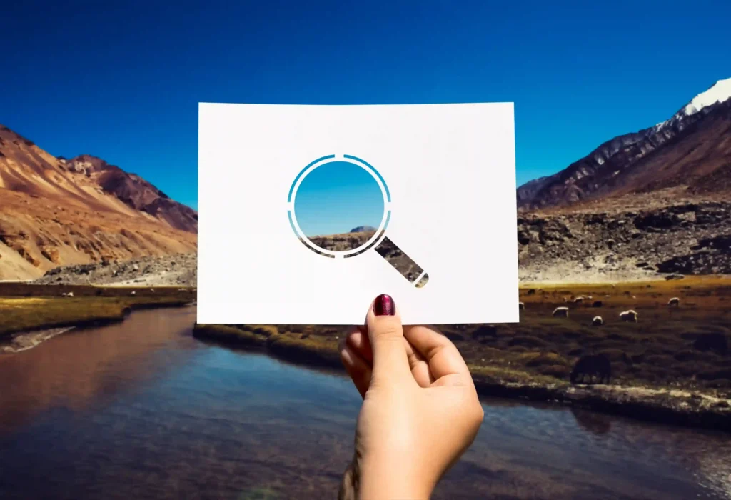 Mão segurando papel com ícone de lupa recortado sobre paisagem ao fundo, representando a pesquisa visual de produto no Mercado Livre para encontrar fornecedores