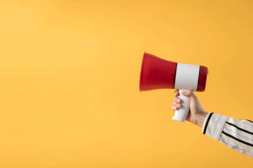 Mão segurando megafone vermelho sobre fundo amarelo, representando a denúncia de plágio de anúncio no Mercado Livre pelos canais corretos