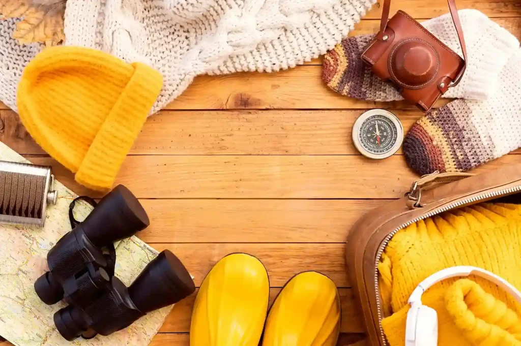 Flatlay de itens de inverno e viagem como gorro, binóculos, botas amarelas, cachecol e mochila sobre madeira, representando as categorias em alta para vender nas férias de julho 2026