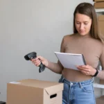 Empreendedora usando leitor de código de barras e caderno para analisar produtos, representando o scanner de catálogo de fornecedor do JoomPulse para encontrar itens lucrativos