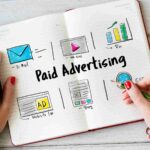 Mãos sobre caderno aberto com esquema desenhado de canais de publicidade paga incluindo e-mail, vídeo, ROI, anúncio em site e blog