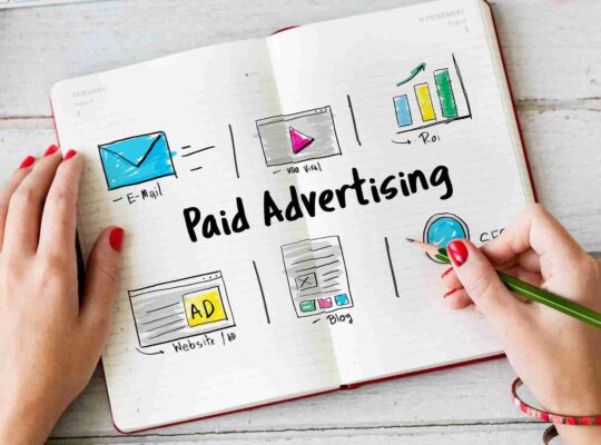 Mãos sobre caderno aberto com esquema desenhado de canais de publicidade paga incluindo e-mail, vídeo, ROI, anúncio em site e blog