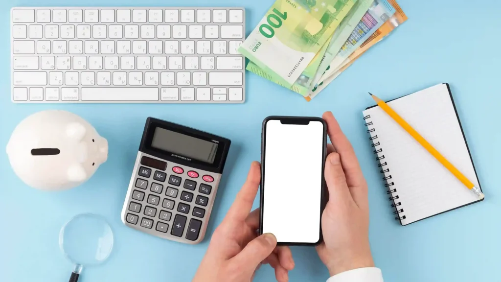 Mãos segurando smartphone com tela em branco ao lado de calculadora, teclado, cofrinho e caderno sobre fundo azul