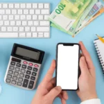 Mãos segurando smartphone com tela em branco ao lado de calculadora, teclado, cofrinho e caderno sobre fundo azul