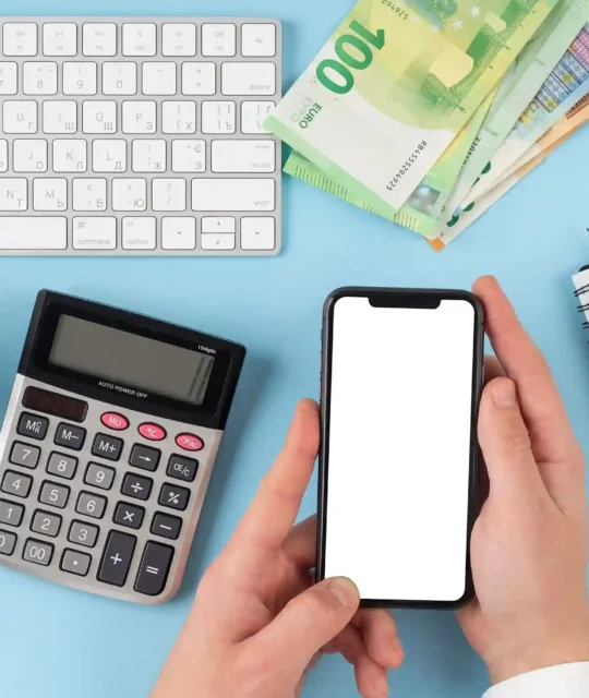 Mãos segurando smartphone com tela em branco ao lado de calculadora, teclado, cofrinho e caderno sobre fundo azul