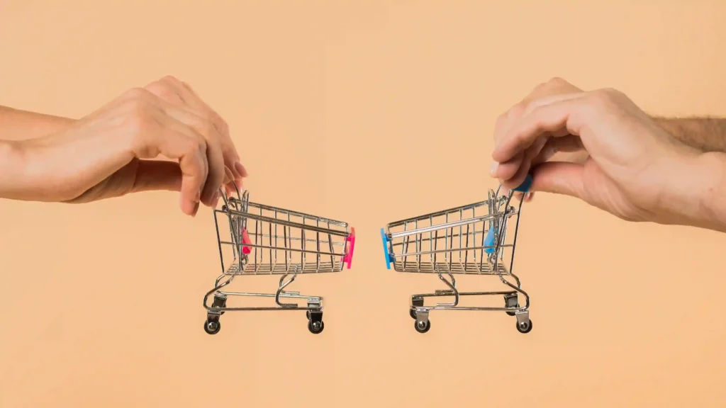 Duas mãos empurrando carrinhos de compras em miniatura um de frente para o outro, sobre fundo bege