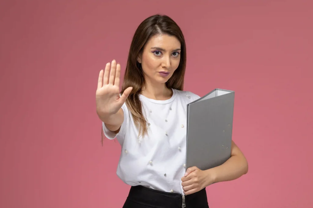 Mulher de blusa branca segurando pasta cinza e fazendo gesto de parada com a mão, fundo rosa