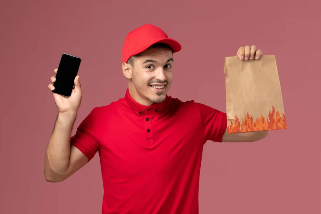 Entregador de uniforme vermelho sorrindo enquanto segura celular e sacola de papel com estampa de chamas, fundo rosa