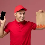 Entregador de uniforme vermelho sorrindo enquanto segura celular e sacola de papel com estampa de chamas, fundo rosa