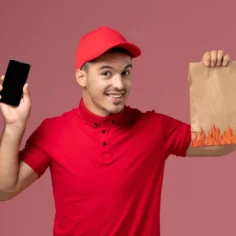 Entregador de uniforme vermelho sorrindo enquanto segura celular e sacola de papel com estampa de chamas, fundo rosa