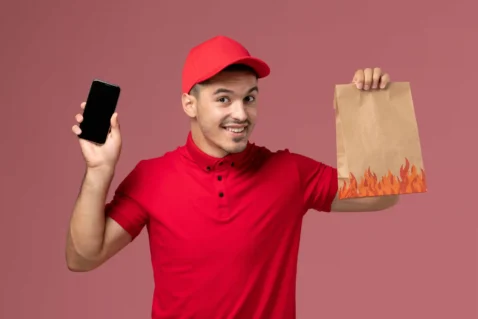 Entregador de uniforme vermelho sorrindo enquanto segura celular e sacola de papel com estampa de chamas, fundo rosa