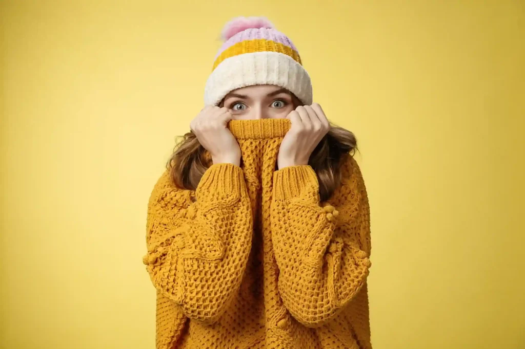 Mulher jovem com touca listrada e suéter amarelo de malha cobrindo parte do rosto com expressão surpresa, fundo amarelo