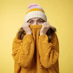 Mulher jovem com touca listrada e suéter amarelo de malha cobrindo parte do rosto com expressão surpresa, fundo amarelo