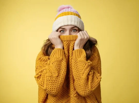 Mulher jovem com touca listrada e suéter amarelo de malha cobrindo parte do rosto com expressão surpresa, fundo amarelo