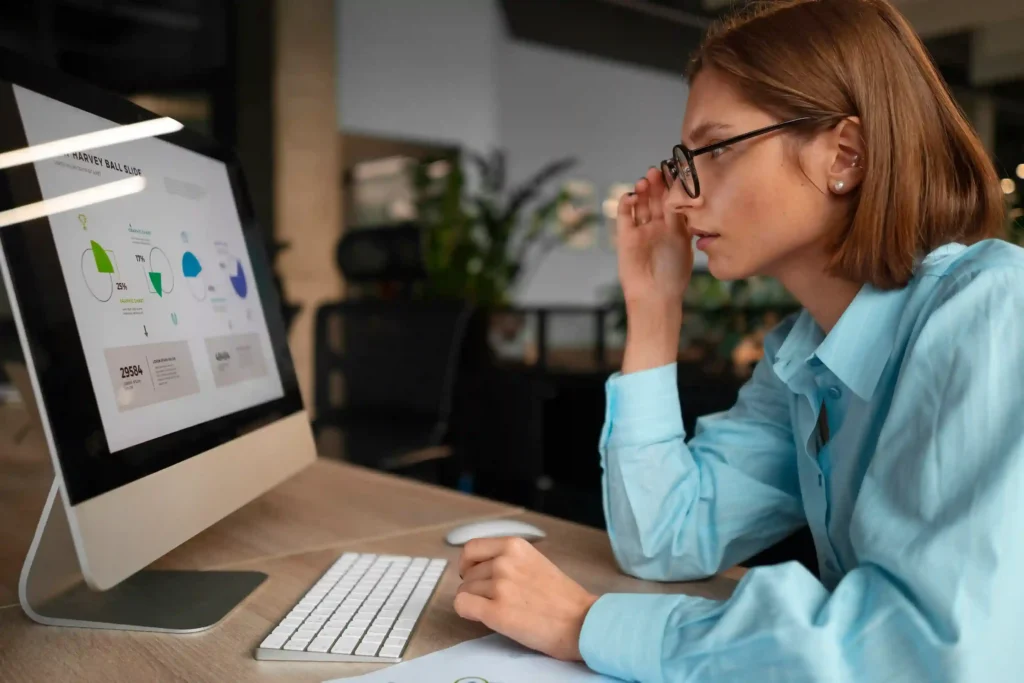 Mulher com óculos e camisa azul observando monitor com gráficos e indicadores em escritório iluminado