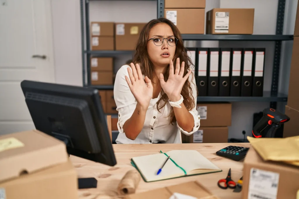 Mulher sentada em uma mesa de escritório, com expressão de preocupação e as mãos levantadas, cercada por caixas, computador e materiais de trabalho