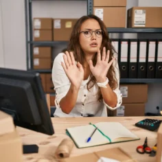 Mulher sentada em uma mesa de escritório, com expressão de preocupação e as mãos levantadas, cercada por caixas, computador e materiais de trabalho