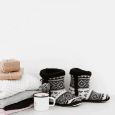 Conjunto de produtos de inverno sobre superfície branca: pilha de mantas dobradas, pantufas de malha, xícara esmaltada e presentes embrulhados