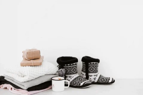 Conjunto de produtos de inverno sobre superfície branca: pilha de mantas dobradas, pantufas de malha, xícara esmaltada e presentes embrulhados