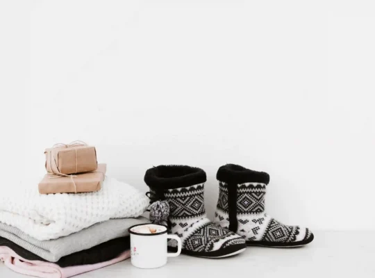 Conjunto de produtos de inverno sobre superfície branca: pilha de mantas dobradas, pantufas de malha, xícara esmaltada e presentes embrulhados