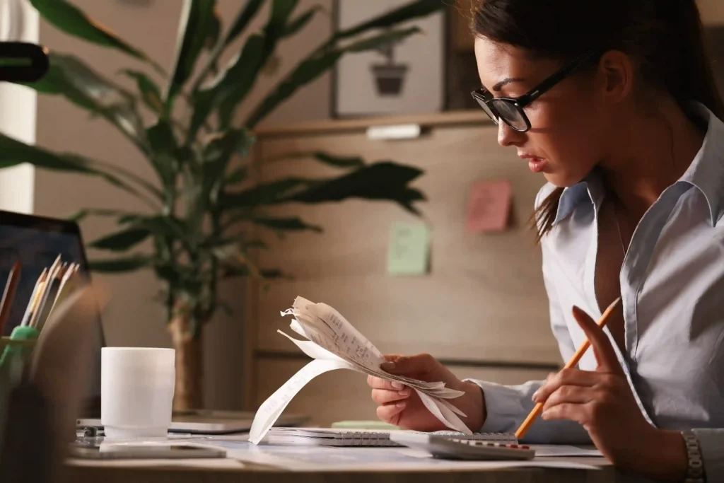 Empreendedora com óculos analisa cupons fiscais em mesa de trabalho com calculadora e caderno
