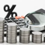 Pilhas decrescentes de moedas ao lado de calculadora, símbolo de porcentagem e lupa sobre fundo branco