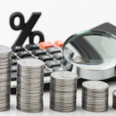 Pilhas decrescentes de moedas ao lado de calculadora, símbolo de porcentagem e lupa sobre fundo branco