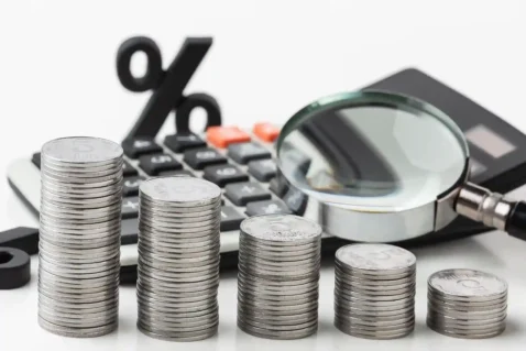 Pilhas decrescentes de moedas ao lado de calculadora, símbolo de porcentagem e lupa sobre fundo branco