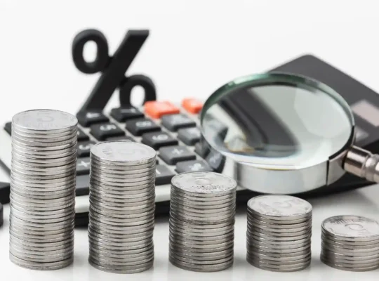 Pilhas decrescentes de moedas ao lado de calculadora, símbolo de porcentagem e lupa sobre fundo branco