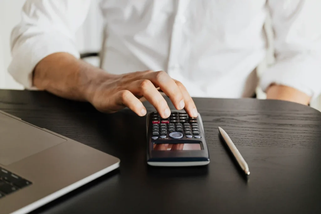 Mão calculando em calculadora científica ao lado de notebook e caneta sobre mesa escura